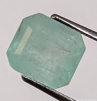 Close up photo of light green emerald cut gemstone held by metal tweezers showing surface and internal inclusions