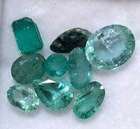 Close-up photo of nine loose genuine emerald gemstones in various shapes and cuts on a white background.