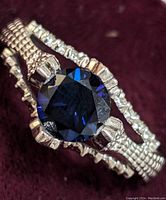 Close-up front view of sterling silver ring with large blue faceted center stone and clear cubic zirconia stones on textured split band