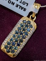 Close-up of sapphire pendant showing three rows of dark blue stones set in gold-tone sterling silver, flanked by clear small stones along edges. Attached to a fine silver chain.