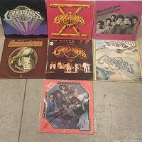 Seven Commodores vinyl album covers displayed in two rows on the floor, showing different designs including a large X, a train, and band photos.