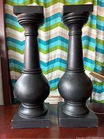 Pair of solid wood black painted pillars with bulbous midsection and square tops standing side-by-side against a colorful curtain background.