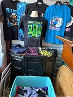 Photo showing multiple graphic t-shirts including Minecraft design and various other event and pop culture graphics displayed on hangers and folded on cabinet.