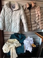 Photo showing two puffy coats, one light grey The North Face and one pewter grey Andrew Marc, hanging side by side above four sweaters in teal blue, off white, cream, and white with decorative elements.