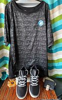 Full view of black moisture-wicking shirt on hanger behind a pair of black and white Adidas high top sneakers, along with wallet and money clip on surface below.