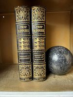 Two vintage Trap Danmark books with black and gold embossed spines labeled Volume I and Volume IV, positioned next to a spherical dark marble weight on a shelf.