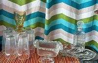 Photo showing one amber drinking glass, eight clear drinking glasses, vintage scalloped dish, Indiana Glass compote bowl, glass decanter, and packing box on a colorful backdrop.
