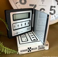 Front view of the digital alarm clock with LCD screen and function buttons, brand and model visible, clock resting on original box.