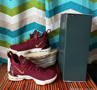 Pair of burgundy LeBron XVI youth sneakers shown with original Nike box, placed on wood surface in front of colorful background curtain.