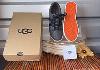 Grey leather UGG youth sneakers, left shoe from top view, right shoe sole with orange bottom visible, next to original UGG box and care booklet.
