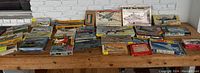Wide view of all model kit boxes laid out on a wooden table showing the collection of 29 aircraft and 1 ship model kits.