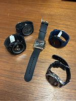 Photo showing all five watches arranged on a wooden surface, some with tags attached on the bands.