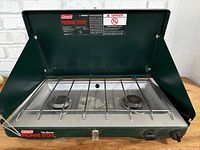 Coleman camping stove with lid open showing two propane burners and metal grate cover.