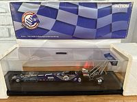 Blue and white dragster model in clear plastic display case with black base, inside styrofoam packaging and blue checkered flag themed cardboard box.