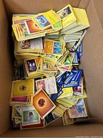 Box filled with a large quantity of mixed Pokémon cards stacked haphazardly showing various card types and colors including energy cards and Pokémon cards.