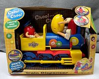 Front view of the M&M's Train Candy Dispenser in original packaging with visible wear and box damage. Shows Red M&M character in conductor seat and Yellow M&M character driving the train, with colorful train body and labels.