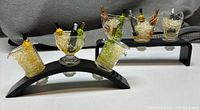 Photo of six miniature acrylic bottle stoppers shaped like ice buckets holding miniature bottles, displayed on a curved black stand.