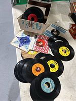 Photo showing multiple 45 RPM vinyl records in paper sleeves and loose records on a table with a decorative box for storage