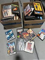 Two cardboard boxes filled with DVDs and VHS tapes, some movie cases laid out in front including Quantum of Solace, Rocky, and others.