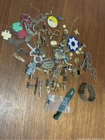 Top view image showing a variety of vintage jewelry and accessory items scattered on wooden surface including cufflinks, pins, brooches, keys, bracelet, religious pendants, tie clips, cameo, and gemstones.