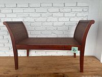 Full side angle view of wooden bench showing woven seat and backrest against white brick wall background.