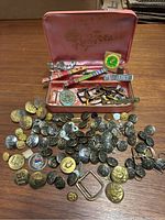 Grouping of various WWII and Cold War military uniform buttons, buckles, pins, and insignia arranged on a wooden surface with a pink lined case in background containing additional items.