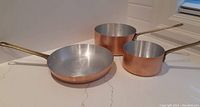 Three copper cookware pieces arranged on countertop showing exteriors and interior tin lining, highlighting brass handles and condition.