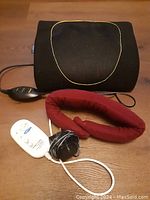 Photo showing black back massager with mesh center and wired controller, alongside red neck massager with wired controller, displayed on wooden floor.