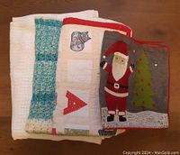 Four folded textile items stacked showing a white blanket, a green and yellow knit blanket, an alphabet quilt with letters, and a Christmas table runner with Santa and tree appliques.