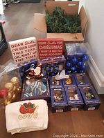 Image showing the overall lot arranged on floor including garland box, plaques, ball ornaments in plastic containers, hand towel, and multiple Kirkland Christmas ornament boxes.