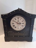 Front view of black wooden clock with round face and Roman numerals, decorative metal studs and panel below the dial.