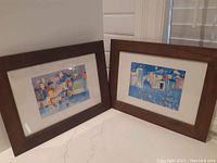 Two framed art prints in brown wooden frames leaning against a wall, showing both prints and frames with scratches.