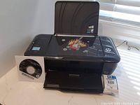 Black HP Deskjet 3000 printer with new HP61 ink cartridge box, installation disc on surface, and power cord connected to printer.