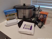 Bravetti crockpot, boxed 2-piece kitchen scale, rice ball maker, dish towel, butter dish, crepe pan, funnels and ice cream scoop all arranged on countertop.