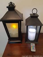 Two black metal lanterns with frosted glass panels and snowflake designs shown side by side on a wooden surface. Larger lantern lit with yellow candle light, smaller lantern lit with white candle light, remote control placed in front.
