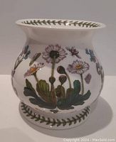 Side view of the ceramic oil burner showing floral botanical design with daisies and leaves in multiple colors on white ceramic base with green leaf border along top and bottom edges.