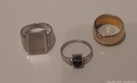 Three rings in a row on flat surface: gold filled thick band, black heart Claddagh silver ring, rectangular top Keyes sterling ring