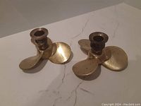 Two solid brass candle holders shaped like three-blade propellers shown on a flat white surface. Each has a candle socket at the center of the propeller.