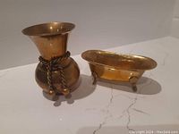 Side angle view of solid brass vase with decorative rope and balls, next to miniature brass tub with claw feet on a marble surface.