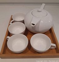 White ceramic teapot with lid and four matching cups on a wooden tray, viewed from above.