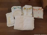 Several piles of folded floral bedsheets in yellow, pink, and blue, plus assorted white lace doilies.