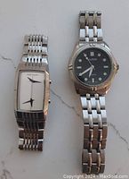 Front view of two men's wristwatches side-by-side on a light surface. Left has a rectangular white dial Rip Curl watch, right a round black dial Alpine watch.