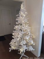 White artificial Christmas tree lit with warm white LED lights, placed indoors with white walls and hardwood floor visible.