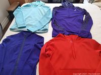 Four women's athletic tops laid out on bed: light blue Under Armour half-zip, purple Adidas pullover, dark blue Lululemon full zip jacket, red Under Armour hoodie.