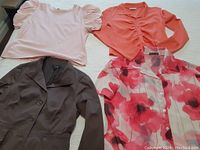 Four women's clothing items laid flat: a light pink short sleeve top with puffed sleeves, a coral ruched long sleeve top, a brown jacket, and a floral oversized button-up shirt with red and pink flowers.
