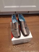 Pair of grey leather women's shoes by Thierry Rabotin on top of a white and red shoe box, shown from a front side angle, displaying the unique pleated cutout design.