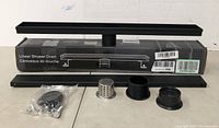 24 inch linear shower drain with black finish, placed on and in front of original packaging box, showing drain channel, metal strainer, two black round fittings, and set of screws with mounting parts sealed in plastic bag.