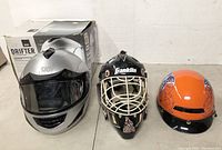Three assorted helmets: silver full-face motorcycle helmet with visor and box, Franklin black ice hockey goalie mask, and orange skateboarding helmet with flame detail