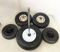 Five assorted wheels arranged in a semi-circle showing different sizes and rim types, including black solid wheels and pneumatic tires with chrome and white rims, one with attached metal shaft.