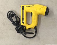 Side view of yellow Stanley electric stapler/nail gun with attached power cord coiled beside it, showing label and trigger.